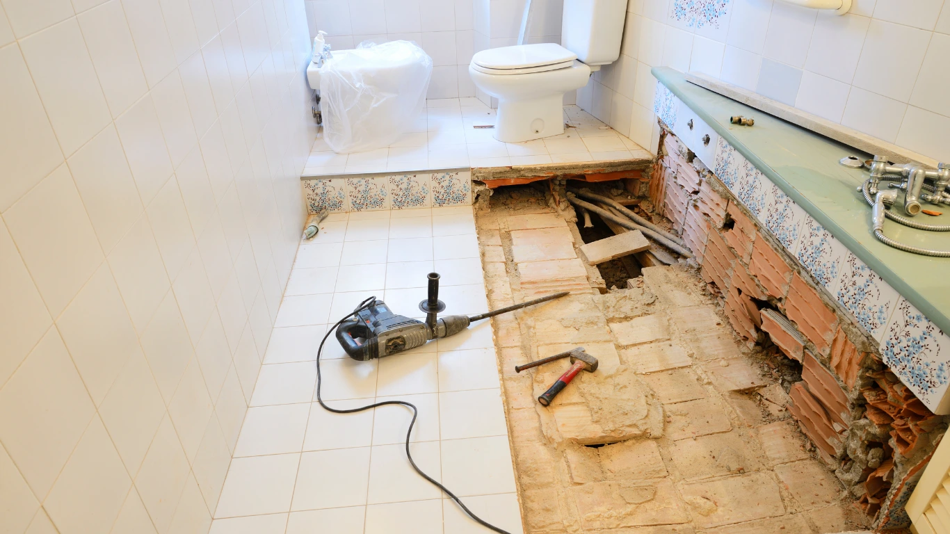 Bathroom mid-renovation with the floor tile torn up to expose the subfloor and plumbing, a rotary hammer and hammer resting on the demolished area beside an old toilet and vanity.