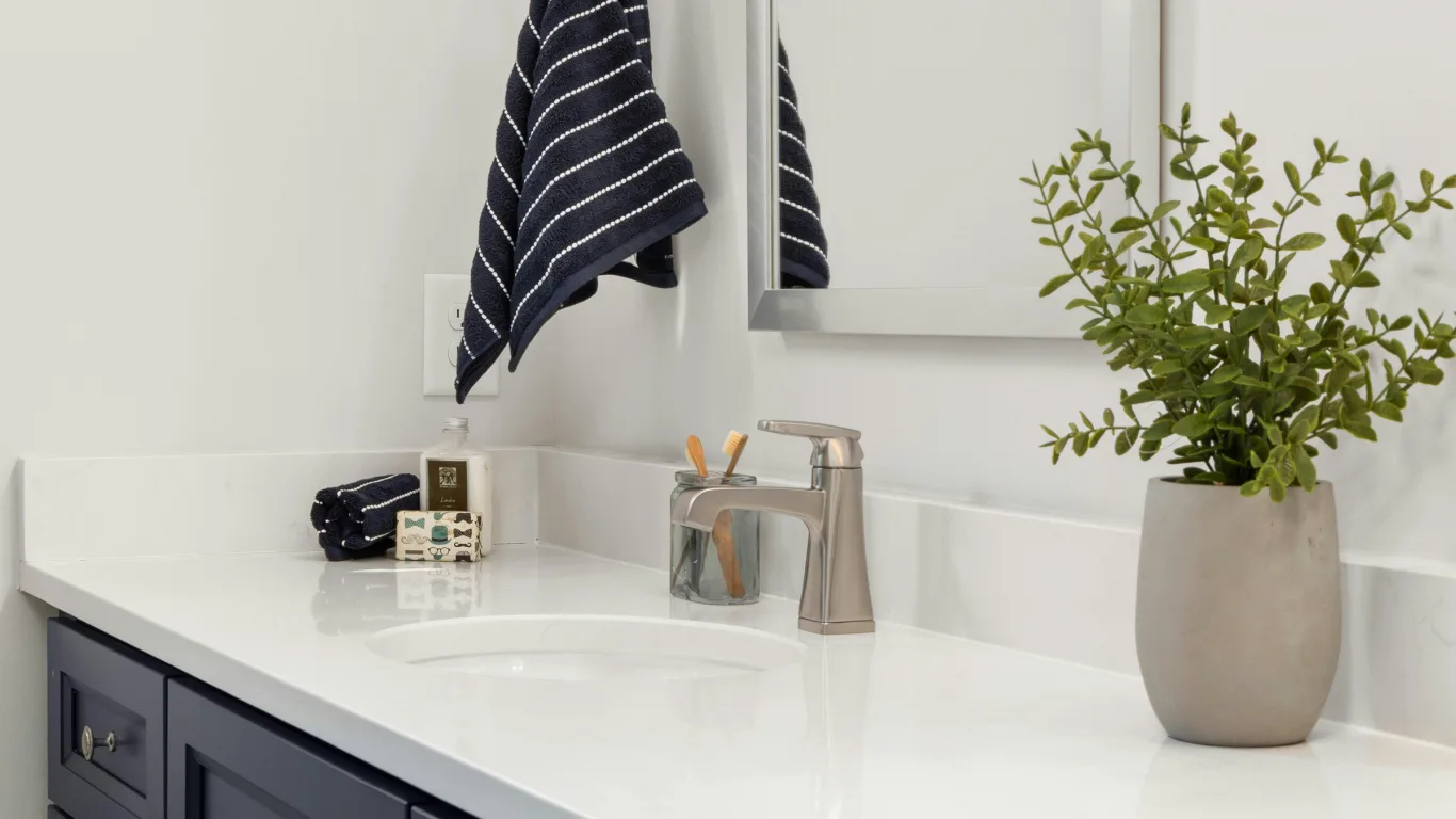 Close-up of a finished bathroom vanity with a white quartz countertop, undermount sink, brushed-nickel faucet, navy cabinets, a striped hand towel, and a potted plant.